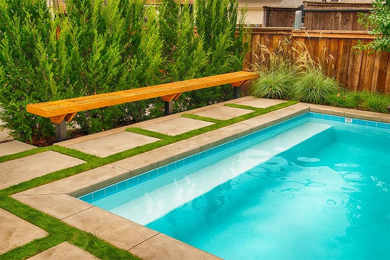 Close up of rectangular swimming pool edge with wooden bench on the deck and landscaping