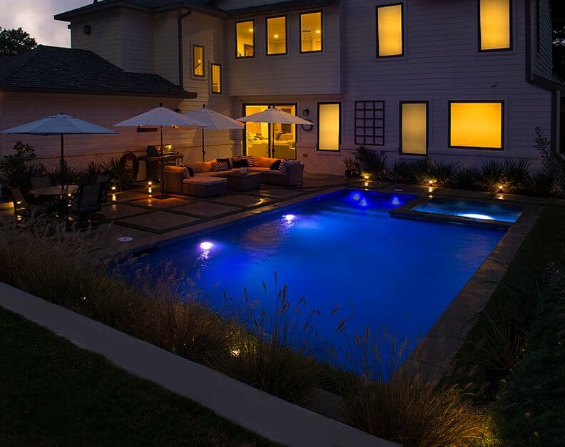 Well lit rectangular backyard swimming pool with nice patio furniture on the porch