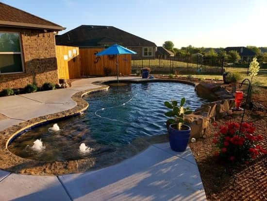 Freeform pool with an umbrella table and three bubblers surrounded by landscaping and an iron fence