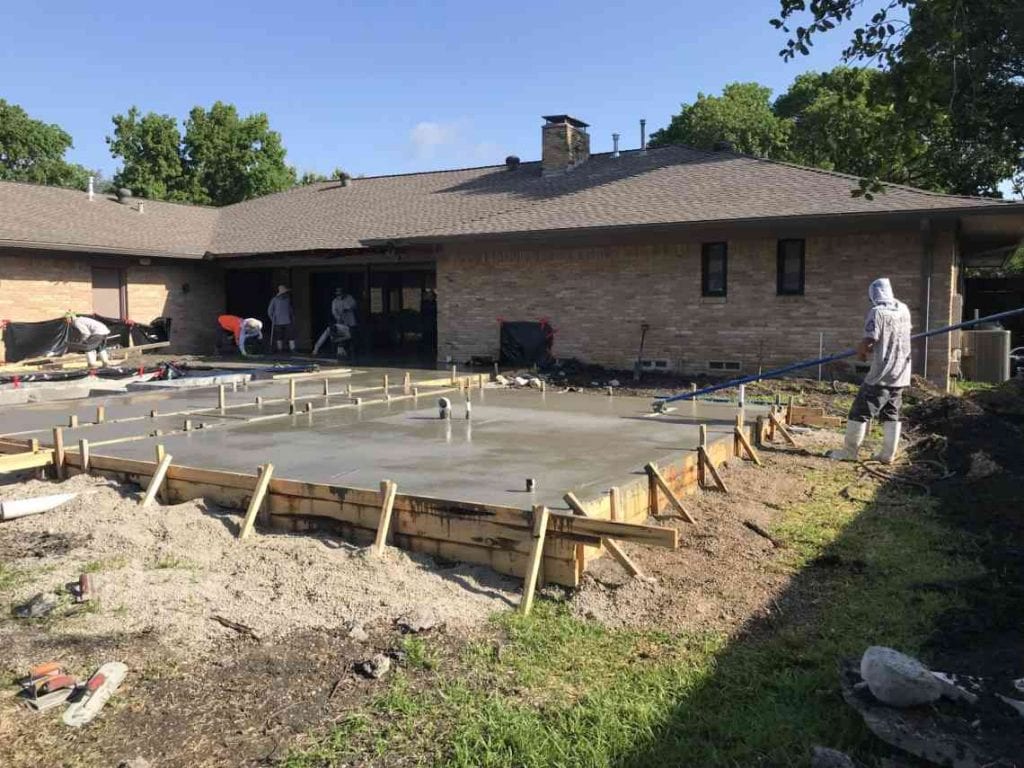 A worker smooths a recently poured concrete deck with a house and some other workers in the background